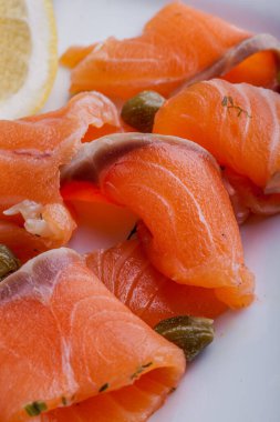 Kaparili somon carpaccio, parmesan dilimleri, zeytinyağı ve baharatlı tost. Yakınlarda bir dilim limon var. Yemekler beyaz tabakta..