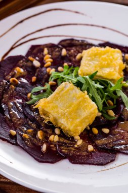 Ballı pancar carpaccio, çam fıstığı, roka yaprağı ve kızarmış tofu. Yemekler yuvarlak, beyaz tabakta. Tabak ahşap bir arka planda duruyor..