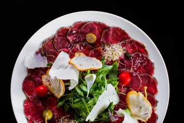 Otlu dana carpaccio. Üstte roka salatası, kapari, parmesan dilimi, karabuğday mikroyeşil, vişneli domates ve buğday ekmeği cipsi karışımı var. Yemekler yuvarlak, beyaz tabakta. Plaka siyah bir arkaplanda duruyor..