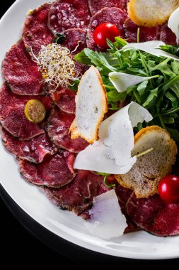 Otlu dana carpaccio. Üstte roka salatası, kapari, parmesan dilimi, karabuğday mikroyeşil, vişneli domates ve buğday ekmeği cipsi karışımı var. Yemekler yuvarlak, beyaz tabakta. Plaka siyah bir arkaplanda duruyor..