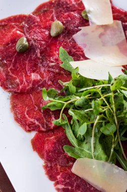 Dana carpaccio roka ve mikroyeşil, parmesan dilimleri, karabiber, balzamik sos ve kapari. Yemek dikdörtgen seramik bir tabakta. Tabak ahşap bir arka planda duruyor..