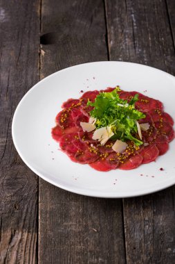 Dijon hardallı dana carpaccio, karışık salata ve parmesan dilimleri. Yemekler yuvarlak, beyaz tabakta. Tabak ahşap bir arka planda duruyor..