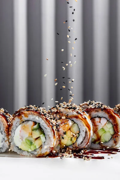 Roll dragon with eel. Roll stuffed with shrimp, avocado, cucumber, Philadelphia cheese wrapped in rice and pieces of smoked eel with unagi sauce, white and black sesame seeds are sprinkled on top. Sushi stands on a white cardboard stand. A stand with