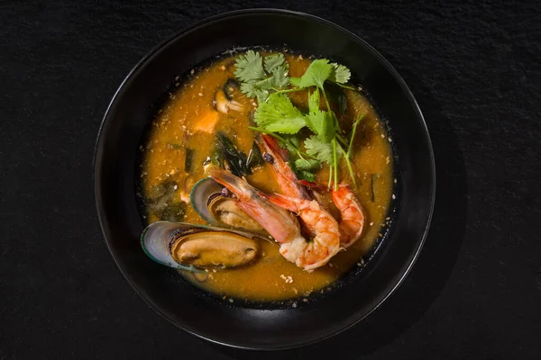 Soup with oysters, shrimps, parsley, sesame seeds and broth in a black plate on a stone black background