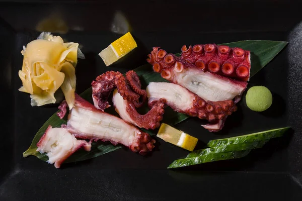 Sliced octopus on a leaf with cucumber, wasabi, lemon and ginger on a black background