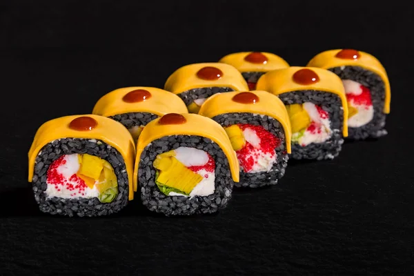 Roll with cheese, sauce, black rice, fruit, squid, tobiko caviar and cream cheese on a black stone background