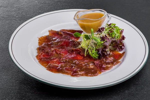 Veal carpaccio with plum sauce, salad mix and gravy boat with mustard sauce. The food is on a round, white plate. The plate stands on a black, metallic background.