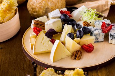 Dört çeşit peynir: dor blue, keçi peyniri, camembert. Yakınlarda cevizler, ahududular, fesleğen yaprakları, üzümler ve mikroyeşil soğan filizleri var. Bal, parmesan peynirinin üstüne dökülür. Yemek yuvarlak, hafif, ahşap bir tahtanın üzerinde duruyor. Yakınlarda.