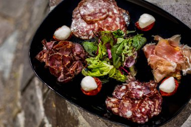 Çeşitli et aperatifleri. Salam, prosciutto, basturma ve baharatlı salam. Yakınlarda küçük mozarella topları olan güneşte kurutulmuş domatesler var. Tabağın ortasında balzamik soslu taze yeşil bir salata var. Yemek siyah, kare bir tabakta. Tabak.