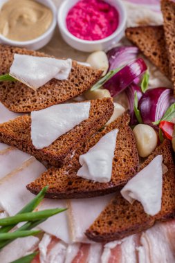 İnce dilimlenmiş domuz yağı ve pastırmalı çavdar tostu, yeşil soğan, turşu, kırmızı biber. Yakında gochitsa ve yabanturplu çay tabakları var. Yemek ahşap bir tabakta. Tabak kırmızı çizgili beyaz bir masa örtüsünün üzerinde duruyor. Yakınlarda ikiye bölünmüş bir soğan yatıyor.