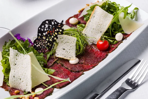 Balzamik soslu dana carpaccio ve otlar. Üstünde roka salatası ve mikroyeşil soğan, buğday ekmeği cipsi, parmesan dilimi, vişneli domates ve Japon mayonezi karışımı. Yemek beyaz, dikdörtgen bir tabakta. Tabak beyaz şeritte.