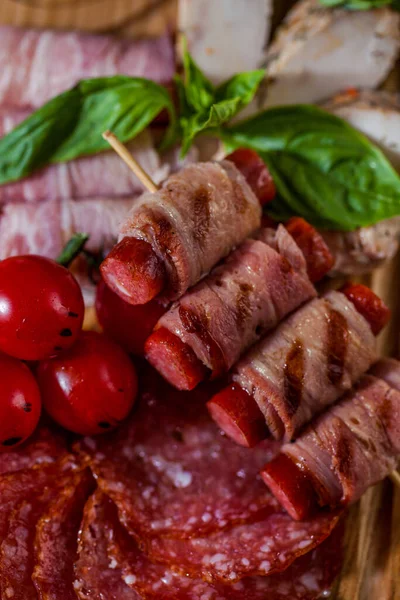 Assorted salami, ham, baked chicken fillet, grilled bacon sausages, lie on a wooden board, on top of basil leaves and a white, ceramic bowl with cranberry sauce on a wooden board. The board lies on a beige tablecloth. Nearby are a fork and a knife, a