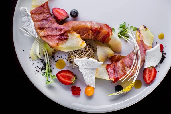 Fresh melon slices wrapped in grilled bacon. Above are the sprouts of microgyne basil. Nearby are strawberries and blueberries, bizet pieces and mango sauce. The food is on a white plate. Black background.