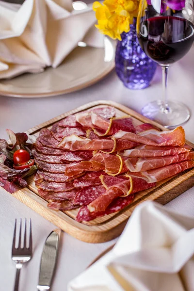 Platter of prosciutto meats. jamon, salami, salami with pepper. Nearby are lettuce leaves with cherry tomatoes cut in half. Assorted lies on a wooden board. The board stands on a white tablecloth, next to it lies a fork and knife, a rag napkin, a cer
