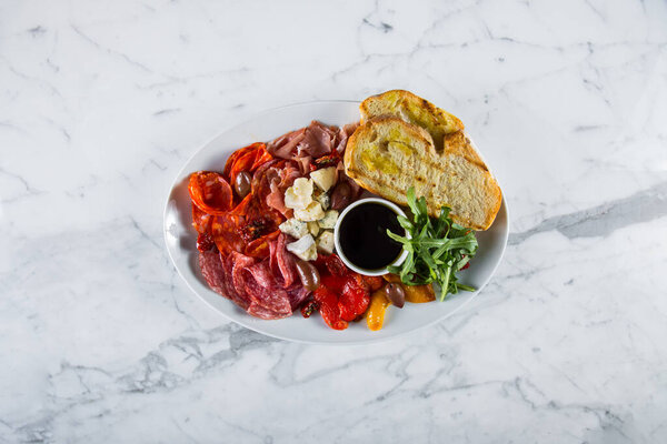 Assorted starters of ham, prosciutto, salami, parmesan, dor blue, arugula leaves, pickled paprika, tomatoes, olives, wheat bread toast. In the center of the plate is a gravy boat with balsamic sauce. The food is on a white, oval plate. Marble backgro