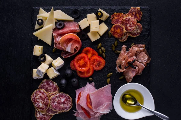 Assorted starters of ham, prosciutto, salami, ham, parmesan, camembert, olives, chopped cherry tomatoes. In the center of the plate is a gravy boat filled with olive oil, inside a teaspoon. The food lies on a black slate plate and stands against a bl