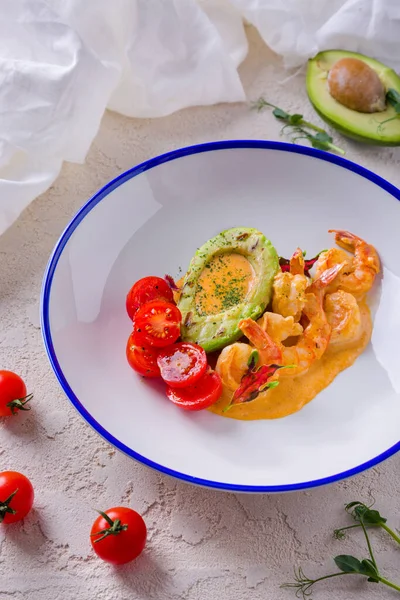 Salad with shrimps, sauce, cherry tomatoes, avocado and arugula in a plate on rough concrete with tomatoes, pea sprouts and half an avocado with a bone and a white cloth on the background