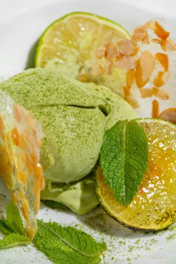 Limonlu ve naneli limonlu dondurma. İki kepçe limonlu dondurma, karamelli kireç parçaları, taze nane yaprakları ve badem buzlu. Üstünde yeşil çay tozu var. Dondurma hafif, derin seramik bir kasede yatıyor. Kase mermer bir ışıkta duruyor.