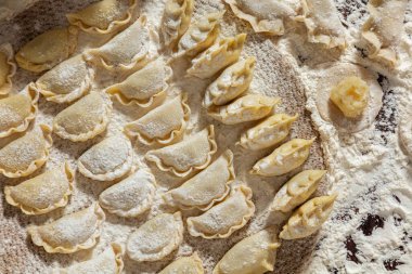 Farklı dolgulu hamur tatlısı. Masanın üzerinde çeşitli şekillerde hamur tatlısı var. Masa un ile kaplı. Yanında hamuru yuvarlamak için bir merdane, unlu bir seramik kase, içinde metal bir kaşık ve keten havlu var. 