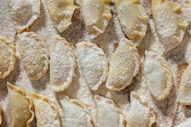 Farklı dolgulu hamur tatlısı. Masanın üzerinde çeşitli şekillerde hamur tatlısı var. Masa un ile kaplı. Yanında hamuru yuvarlamak için bir merdane, unlu bir seramik kase, içinde metal bir kaşık ve keten havlu var. 