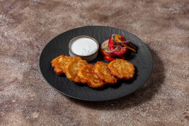 Kızartma tavasında tereyağlı ızgara patates gözlemesi yanında kırmızı soğan, paprika ve domates salatası, ayçiçeği yağı ve ince doğranmış maydanoz serpiştirilmiş. Yakınlarda ekşi kremalı bir seramik kase var. Yiyecekler siyah renkte.