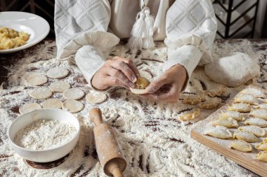 Hamur tatlısı yapma süreci. Kadın beyaz, geleneksel Ukrayna vyshyvanka 'sı ve kahverengi bir etek giyiyor. Elleriyle masada hamur tatlısı yapıyor. Patates püresini yuvarlak hamur parçalarına sarar. Masa un ile kaplı. Yaklaş.