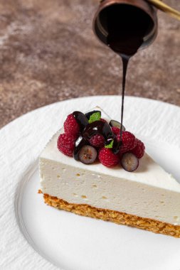 Kuş sütlü kek, ahududu ve böğürtlenli kurabiye ve fındık. Sıcak, siyah çikolata tatlının üstüne dökülüyor ve ezilmiş fındıklar serpiştiriliyor. Pasta beyaz, yuvarlak, seramik bir tabağın üzerinde duruyor. Tabak siyah bir kumaşın üzerinde duruyor. 