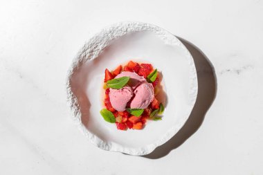 Çilekli dondurma, çilekli ve taze naneli. Üç kepçe çilekli dondurma ince doğranmış çilek yastığının üzerinde duruyor ve üzerinde taze nane yaprakları var. Dondurma ve böğürtlen derin, hafif seramik bir tabakta yatar. Tabak üzerinde duruyor.