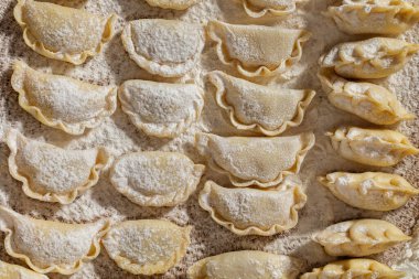 Farklı dolgulu hamur tatlısı. Masanın üzerinde çeşitli şekillerde hamur tatlısı var. Masa un ile kaplı. Yanında hamuru yuvarlamak için bir merdane, unlu bir seramik kase, içinde metal bir kaşık ve keten havlu var. 