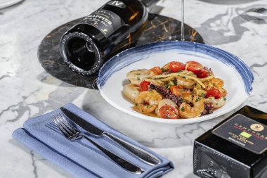 Deniz mahsullü makarna. Deniz tarağı, ahtapot, karides ve kremalı bitki soslu kiraz domatesli ev yapımı makarna. Makarna yuvarlak, hafif seramik tabakta yatıyor. Tabak hafif bir mermer masanın üzerinde duruyor. Yanında bir bardak Prosecco, keten peçetede çatal bıçak takımı.