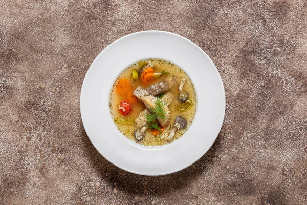 Fish soup Ukha from sturgeon. In a white, round, ceramic plate, there are two pieces of sturgeon fillet, chopped carrots and fish broth. Nearby is a small, white, ceramic bowl with finely chopped dill and parsley. The dishes stand on a dark, fabric b