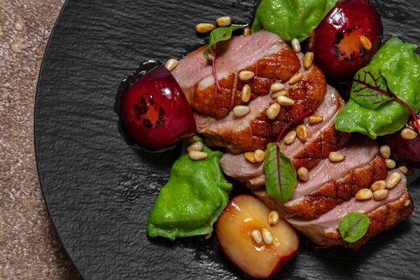 Duck breast in plum sauce, grilled and cut into portions. Topped with pine nuts and lettuce leaves. Nearby are basil ravioli with mashed potatoes and porcini mushrooms. Plum sauce and caramelized plums lie side by side. The food lies on a black, slat