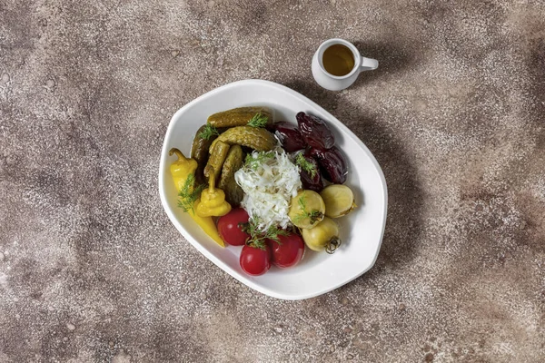 A plate of pickled vegetables. The plate contains pickled red tomatoes, pickled green tomatoes, pickled cucumbers, sauerkraut, pickled peppers and prunes. Pickles lie in an oval, white, ceramic plate. The plate stands on a fabric background, coffee c