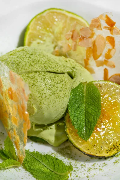 Lime ice cream with lime wedges and mint. Two scoops of lime ice cream with caramelized lime chunks, fresh mint leaves and almond ice cubes. Topped with green tea powder. Ice cream lies in a light, deep ceramic bowl. The bowl stands on a marble light