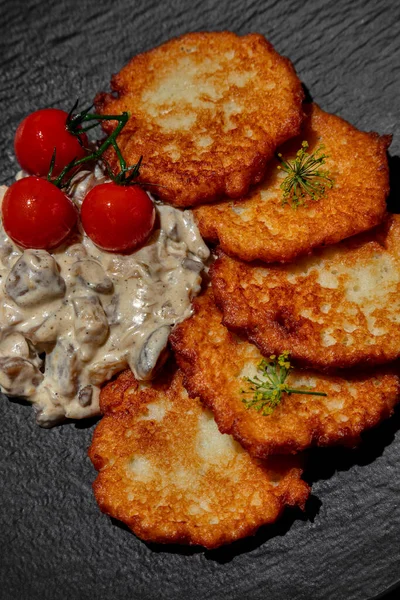 Potato pancakes with dill seeds, cherry tomatoes and mushrooms in sauce in a plate on a wooden background