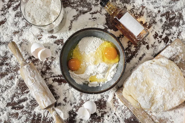 Ingredients for cooking Ukrainian dish dumplings. Flour is scattered on the kitchen table, eggshells, various glass bottles and jars of sauces and oils lie. There is a wooden board with dough, there is a rolling pin for rolling out the dough, there i