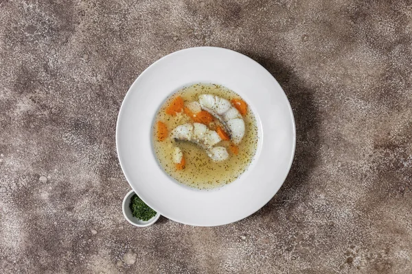 Fish soup Ukha from sturgeon. In a white, round, ceramic plate, there are two pieces of sturgeon fillet, chopped carrots and fish broth. Nearby is a small, white, ceramic bowl with finely chopped dill and parsley. The dishes stand on a dark, fabric b