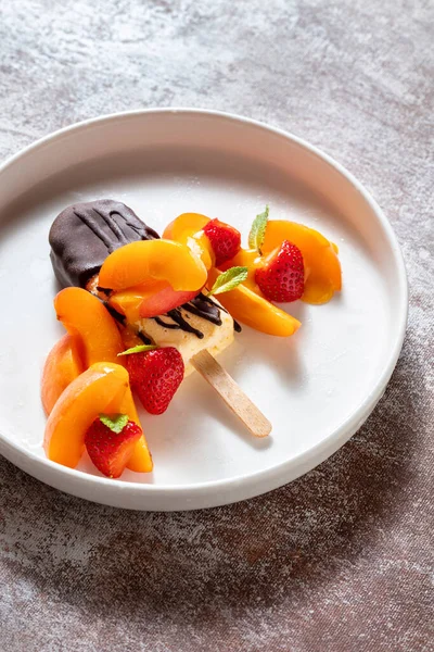 Ice cream on a stick in dark chocolate with peaches, strawberries, mint on a plate