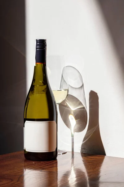 Bottle of white wine on the table, in the sunlight. Nearby is a glass of white wine. The bottle and glass stand on a varnished table and are reflected. A dark brown table stands against a white wall with Ukrainian ornaments.