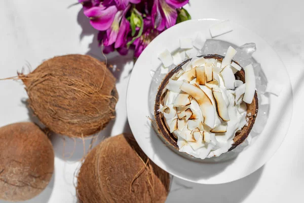 Hindistan cevizli dondurma ve hindistan cevizi. Hindistan cevizli iki kepçe dondurma hindistan cevizli kabuğun içinde, ince doğranmış hindistan cevizli et üzerine serpilmiş. Dondurmalı kabuğun yarısı beyaz bir seramik kasede buzdan bir tepenin üzerinde yatıyor. Kase bir 