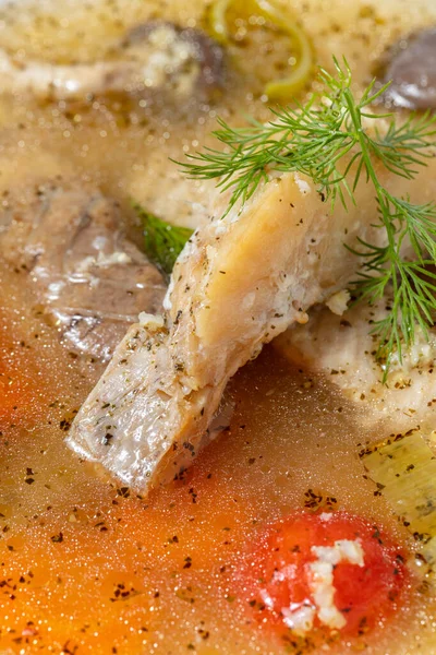 Fish soup Ukha from sturgeon. In a white, round, ceramic plate, there are two pieces of sturgeon fillet, chopped carrots and fish broth. Nearby is a small, white, ceramic bowl with finely chopped dill and parsley. The dishes stand on a dark, fabric b