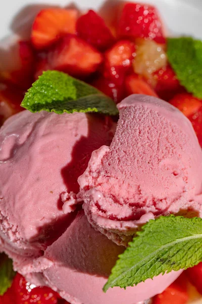 Strawberry ice cream with strawberries and fresh mint. Three scoops of strawberry ice cream lie on a pillow of finely chopped strawberries, with fresh mint leaves on top. Ice cream and berries lie in a deep, light ceramic plate. The plate stands on a