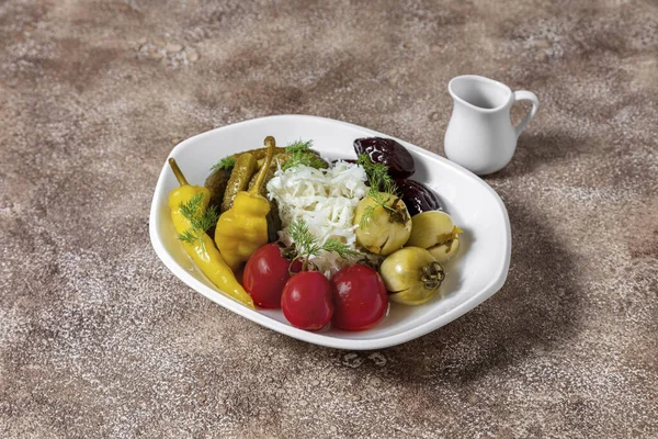 A plate of pickled vegetables. The plate contains pickled red tomatoes, pickled green tomatoes, pickled cucumbers, sauerkraut, pickled peppers and prunes. Pickles lie in an oval, white, ceramic plate. The plate stands on a fabric background, coffee c