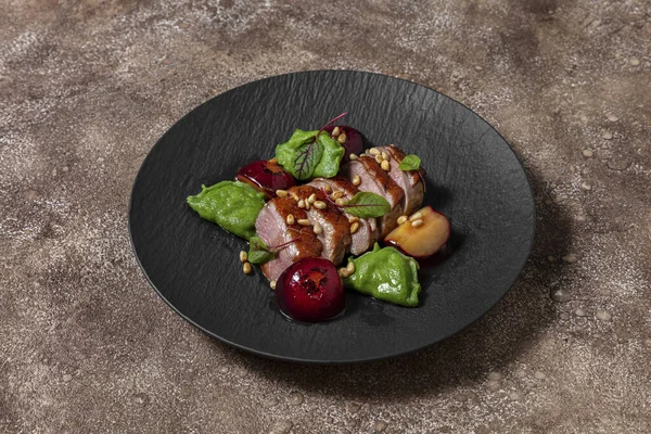 Duck breast in plum sauce, grilled and cut into portions. Topped with pine nuts and lettuce leaves. Nearby are basil ravioli with mashed potatoes and porcini mushrooms. Plum sauce and caramelized plums lie side by side. The food lies on a black, slat