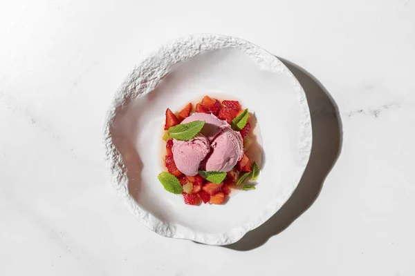 Strawberry ice cream with strawberries and fresh mint. Three scoops of strawberry ice cream lie on a pillow of finely chopped strawberries, with fresh mint leaves on top. Ice cream and berries lie in a deep, light ceramic plate. The plate stands on a