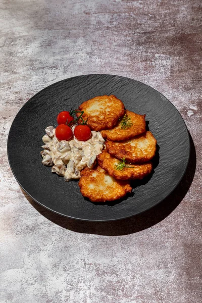 Potato pancakes with dill seeds, cherry tomatoes and mushrooms in sauce in a plate on a gray background