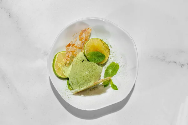 Lime ice cream with lime wedges and mint. Two scoops of lime ice cream with caramelized lime chunks, fresh mint leaves and almond ice cubes. Topped with green tea powder. Ice cream lies in a light, deep ceramic bowl. The bowl stands on a marble light