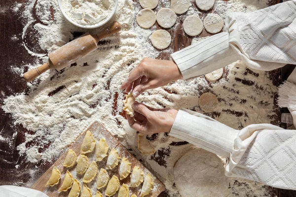 The process of making dumplings. The woman is dressed in a white, traditional Ukrainian vyshyvanka and a brown skirt. She makes dumplings on the table with her hands. Wraps mashed potatoes in round dough pieces. The table is covered with flour. Nearb