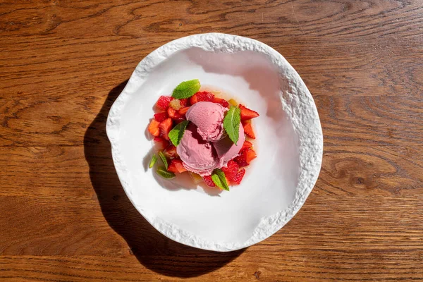 Strawberry ice cream with strawberries and fresh mint. Three scoops of strawberry ice cream lie on a pillow of finely chopped strawberries, with fresh mint leaves on top. Ice cream and berries lie in a deep, light ceramic plate. The plate stands on a