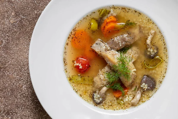 Fish soup Ukha from sturgeon. In a white, round, ceramic plate, there are two pieces of sturgeon fillet, chopped carrots and fish broth. Nearby is a small, white, ceramic bowl with finely chopped dill and parsley. The dishes stand on a dark, fabric b